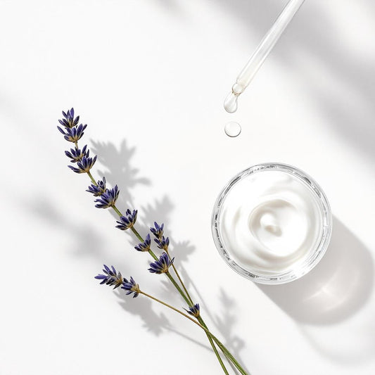 A minimal skincare scene featuring a glass jar of white cream, a dropper releasing a clear droplet above it, and a sprig of lavender on a bright white background.